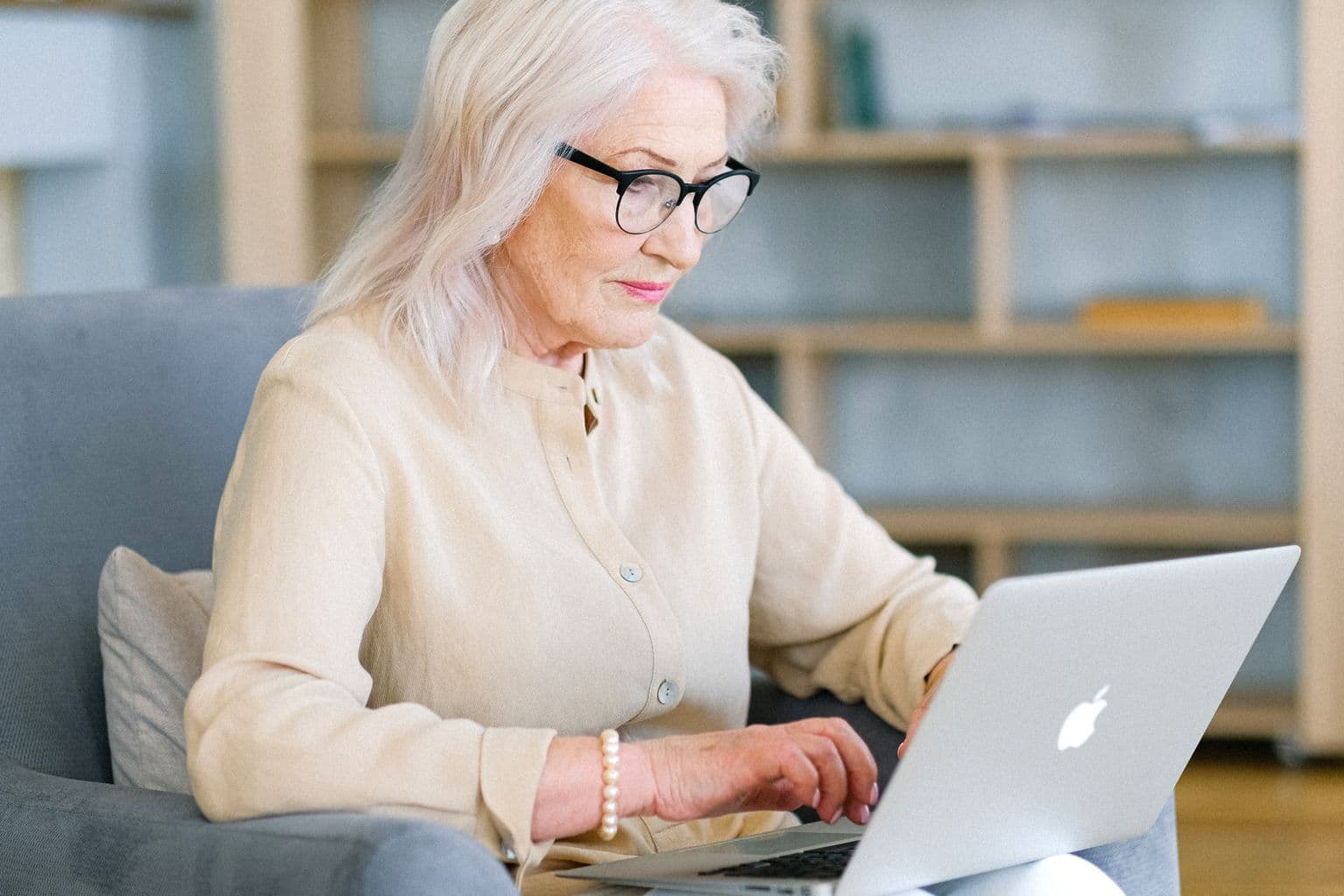 An older woman wearing glasses and using a laptop in an armchair.